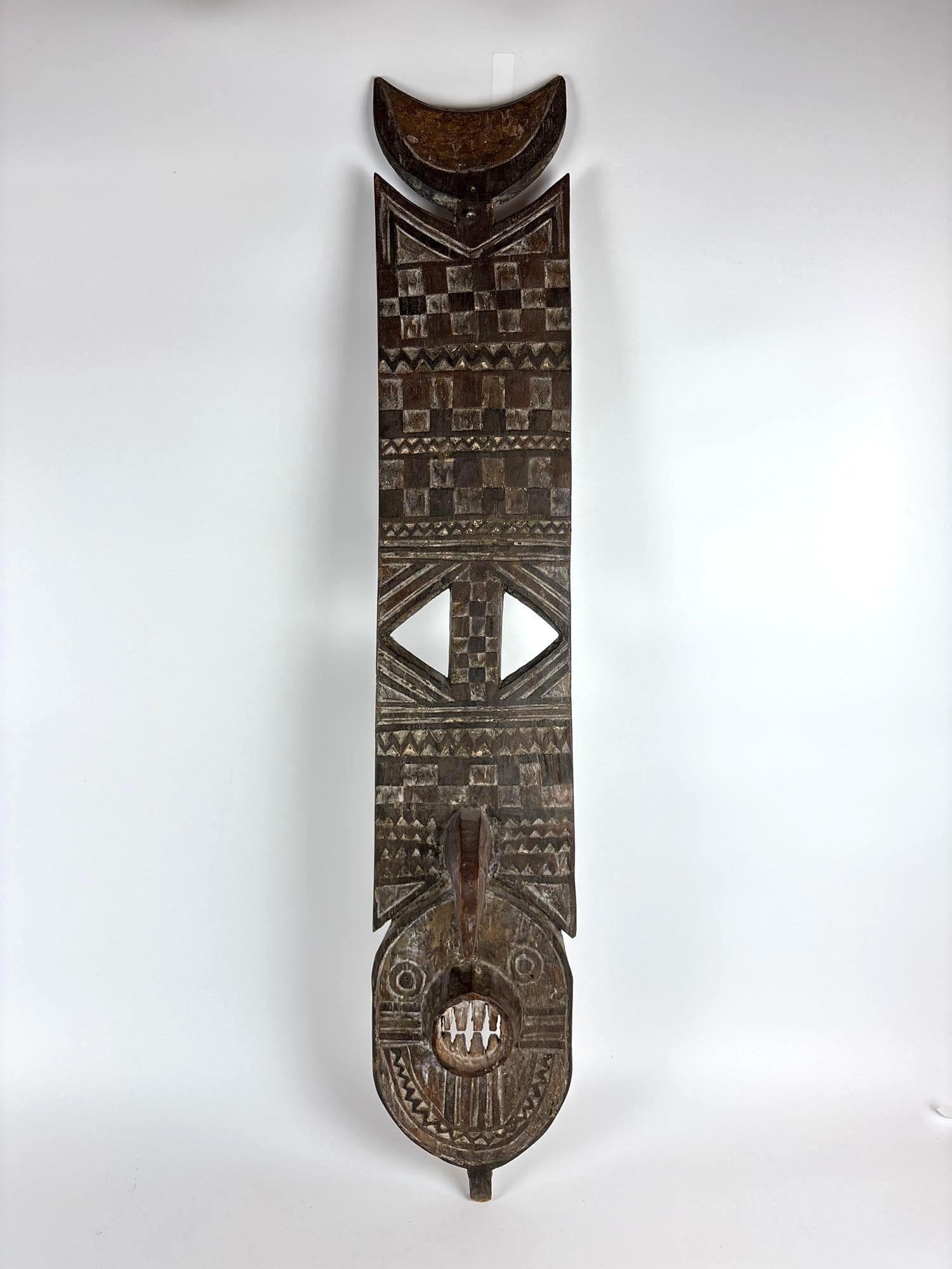 MOSSI PEOPLE, KARANGA MASK: A tall, narrow, carved wooden mask with an oval face and a tall plank-like superstructure decorated with incised geometric patterns and a crescent moon shape at the very top. Dimensions: 44" X 7" X 6"