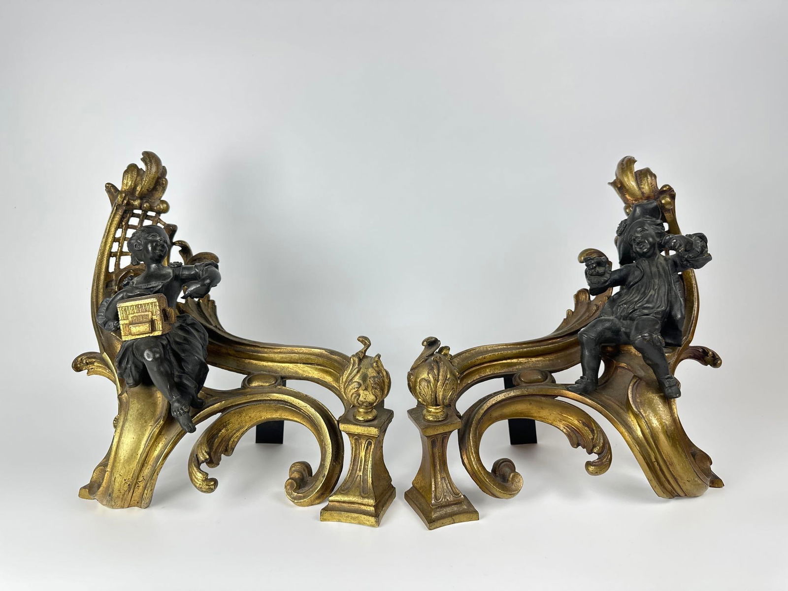 PAIR OF LOUIS XV STYLE FIGURAL PARCEL GILT BRONZE CHENETS: A pair of gilt and patinated bronze fireplace accessories in the Louis XV style, featuring scrolling rococo forms and opposing seated figures, with one holding a box. Dimensions: 12"T x 12"W