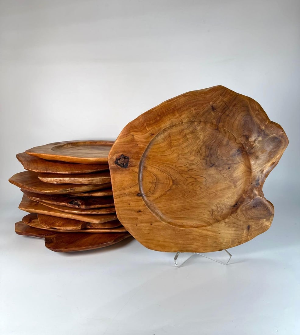 ENRICO SET OF 12 LIVE EDGE HARDWOOD CHARGER PLATES: A set of 12 live edge hardwood trays featuring a smooth, recessed circular center, and visible wood grain patterns and knots in a warm, medium brown tone. Various Dimensions: from approx. 14"-16&