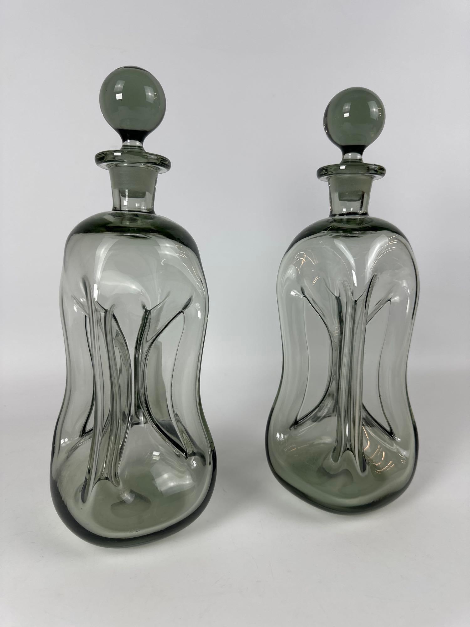 KLUK KLUK DECANTER SET OF TWO JACOB E. BANG SMOKED ART GLASS DECANTERS FOR HOLMEGAARD: These are two Kluk Kluk decanters in smoked art glass designed by Jacob E. Bang for Holmegaard. Dimensions: 13”H x 5”W x 5”D Maker: Holmegaard Provenance: The Estate of Joseph Roda