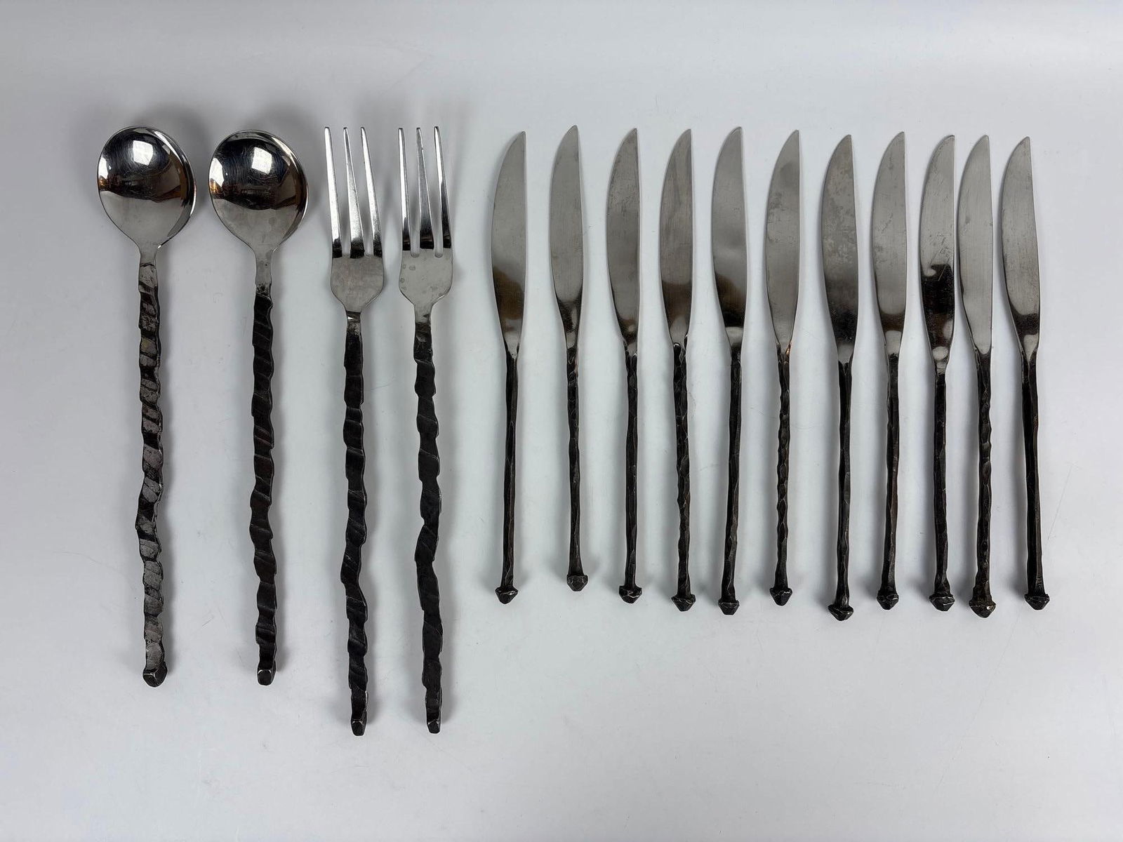 15 PIECE COLLECTION OF MICHAEL ARAM "IRON AGE" STAINLESS STEEL FLATWARE: A unique collection of handmade stainless steel flatware consisting of two serving spoons, two serving forks, and eleven knives, all with distinctive, slender oxidized black handles. Largest Dimensio