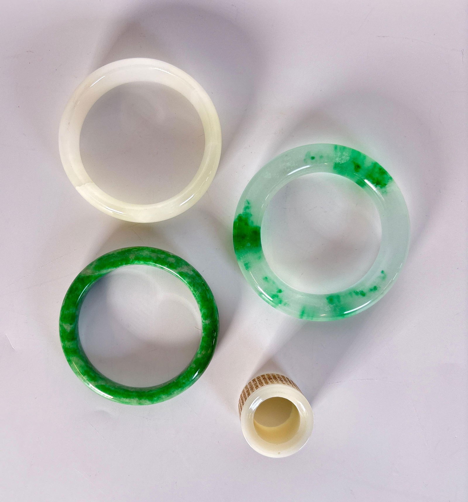 GROUP OF THREE JADE / JADEITE BANGLES AND ONE ARCHER'S RING: This lot comprises four pieces of carved and polished stone: three circular bangles in varied shades of white, celadon, and mottled green jadeite or jade, and one thick, carved white jade archer's