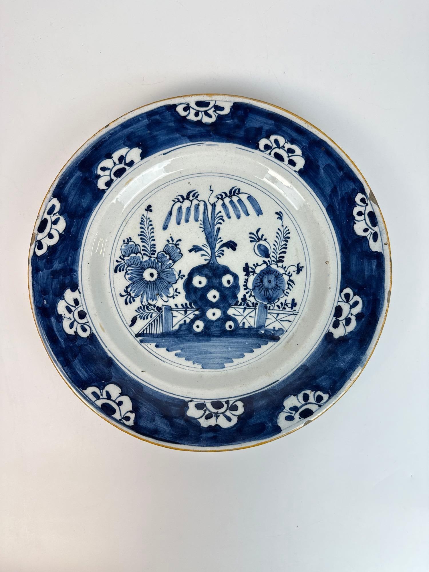 18TH C. DUTCH DELFT BLUE & WHITE CHARGER – EXAGGERATED FLOWERS: Large tin-glazed earthenware charger with ochre rim, hand-painted scrolling foliate motifs blue floral border, and central design of stylized oversized blossoms flanking a tree and fence. Approximat