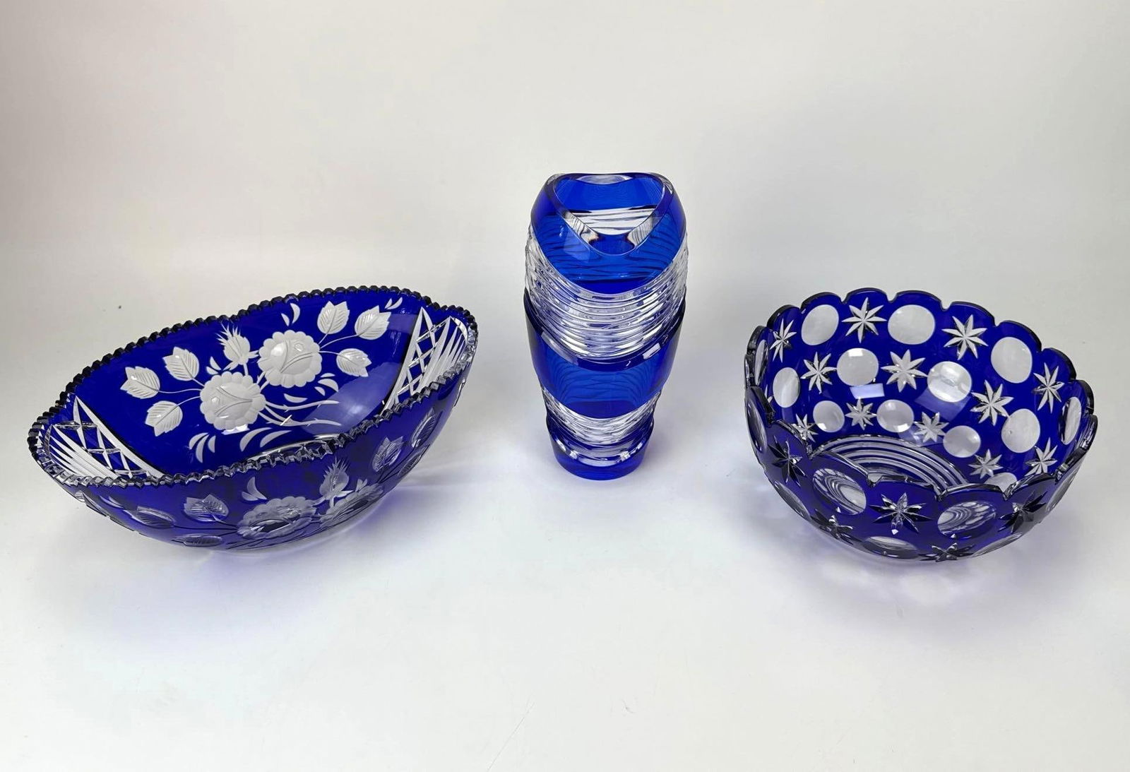 BOHEMIAN COBALT BLUE CUT TO CLEAR CRYSTAL VESSEL TRIO: A three-piece collection of vintage cobalt blue cut-to-clear crystal including one vase and two decorative bowls with floral and geometric patterns. Round bowl is signed at the bottom but indecipherab