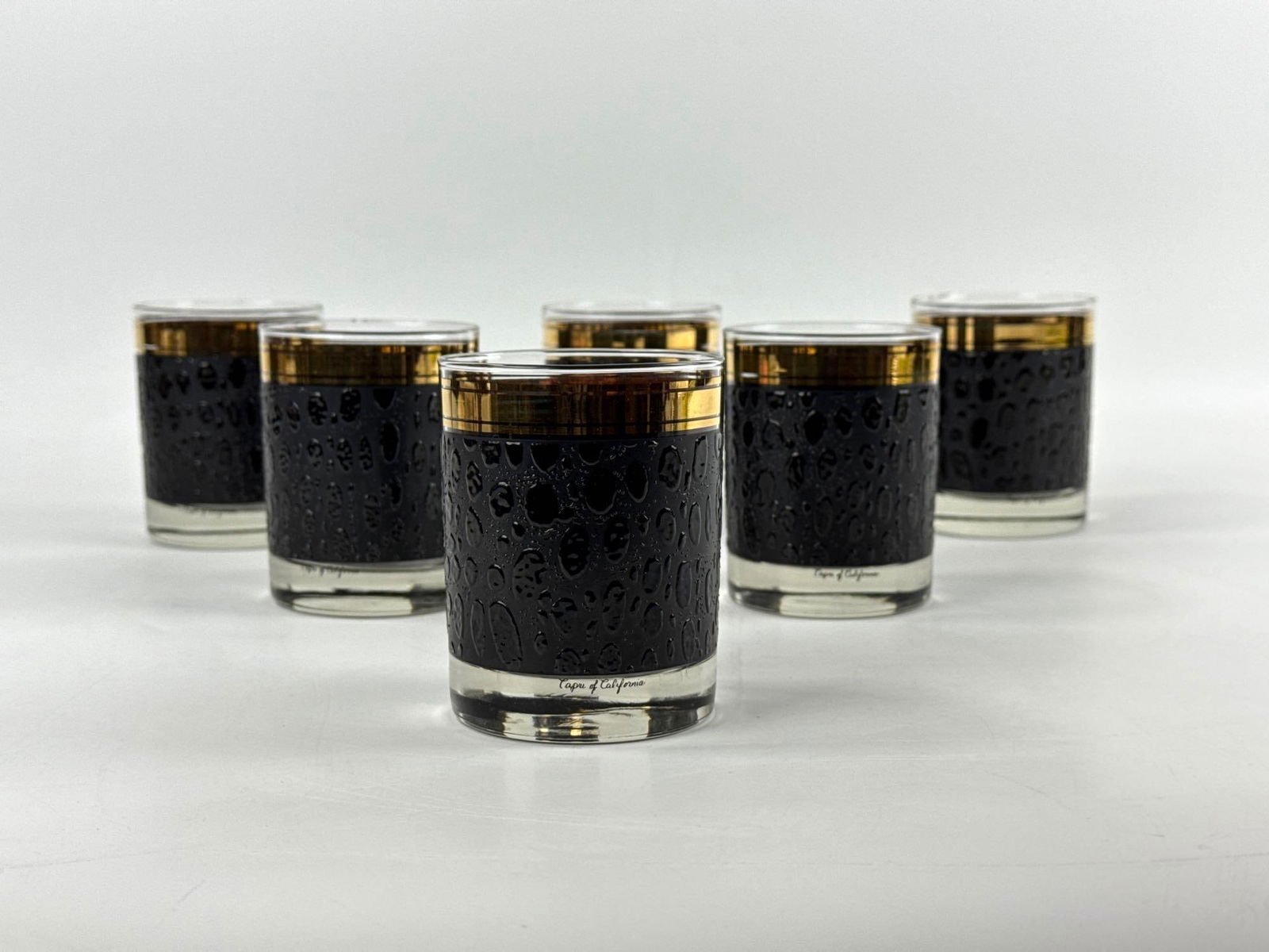 SET OF 6 VINTAGE CAPRI OF CALIFORNIA LEOPARD LOWBALL GLASSES: Mid-century modern whiskey glasses with a tactile black embossed leopard print design and wide 22kt gold rims. Approximate Dimensions: 4 1/8”h x 3 1/4dia Artist or Maker: Capri of California Pro