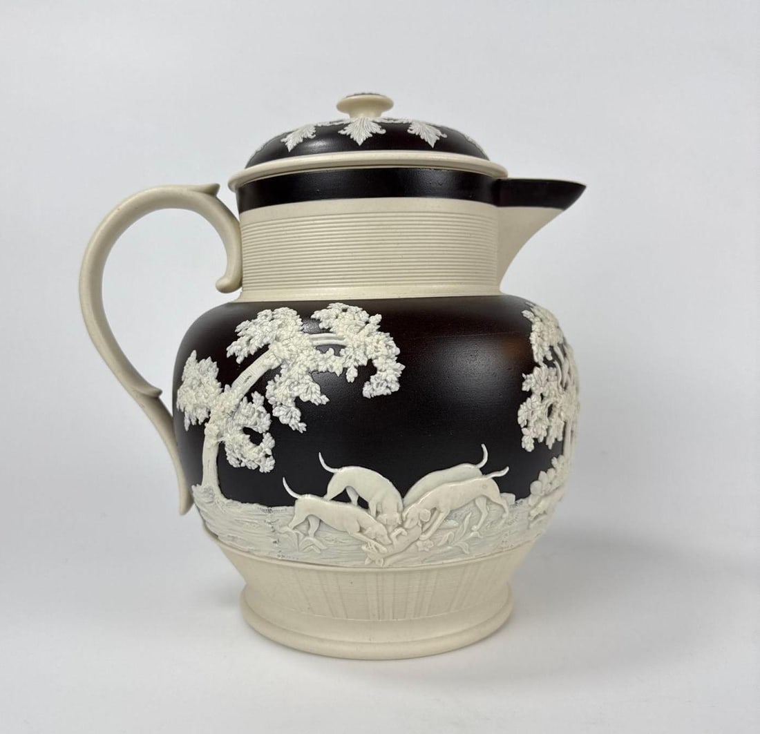 ANTIQUE BLACK AND WHITE JASPERWARE HUNT SCENE PITCHER WITH LID: *Black stoneware pitcher with white sprigged relief decoration showing hunting dogs in pursuit of a stag amidst woodland trees Approximate Dimensions: 10”h x 9.75”w x 7.5”d Artist or