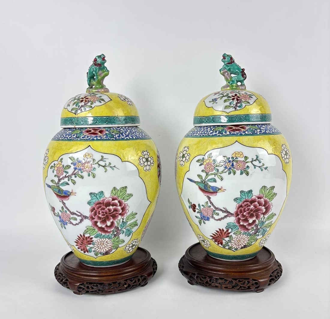 PAIR OF FRENCH YELLOW PORCELAIN COVERED JARS WITH FOO LION FINIALS AND CARVED TEAK BASES: *Twin hand-painted Famille Rose Chinoiserie lidded ceramic baluster form vessels and matching lids topped with a foo dog finial on wood openwork stands. One with previous restoration (see photos). Ap