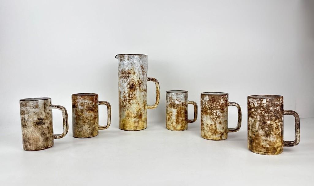 VINTAGE SIGNED ALEXANDRE KOSTANDA VALLAURIS STONEWARE PITCHER AND MUGS: *Hand-thrown French studio pottery set consisting of one tall cylindrical pitcher and five matching mugs with rustic earth-toned glazes and distinctive spotted textures Approximate Dimensions: Pitcher