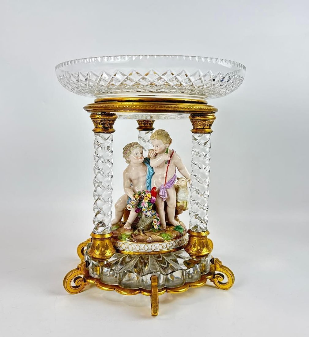 MEISSEN PORCELAIN AND BACCARAT CRYSTAL FIGURAL CENTERPIECE: Antique centerpiece with Meissen porcelain cherub group housed within a Baccarat cut crystal and gilt bronze architectural stand supported by four spiral columns. Approximate Dimensions: 9.75”h