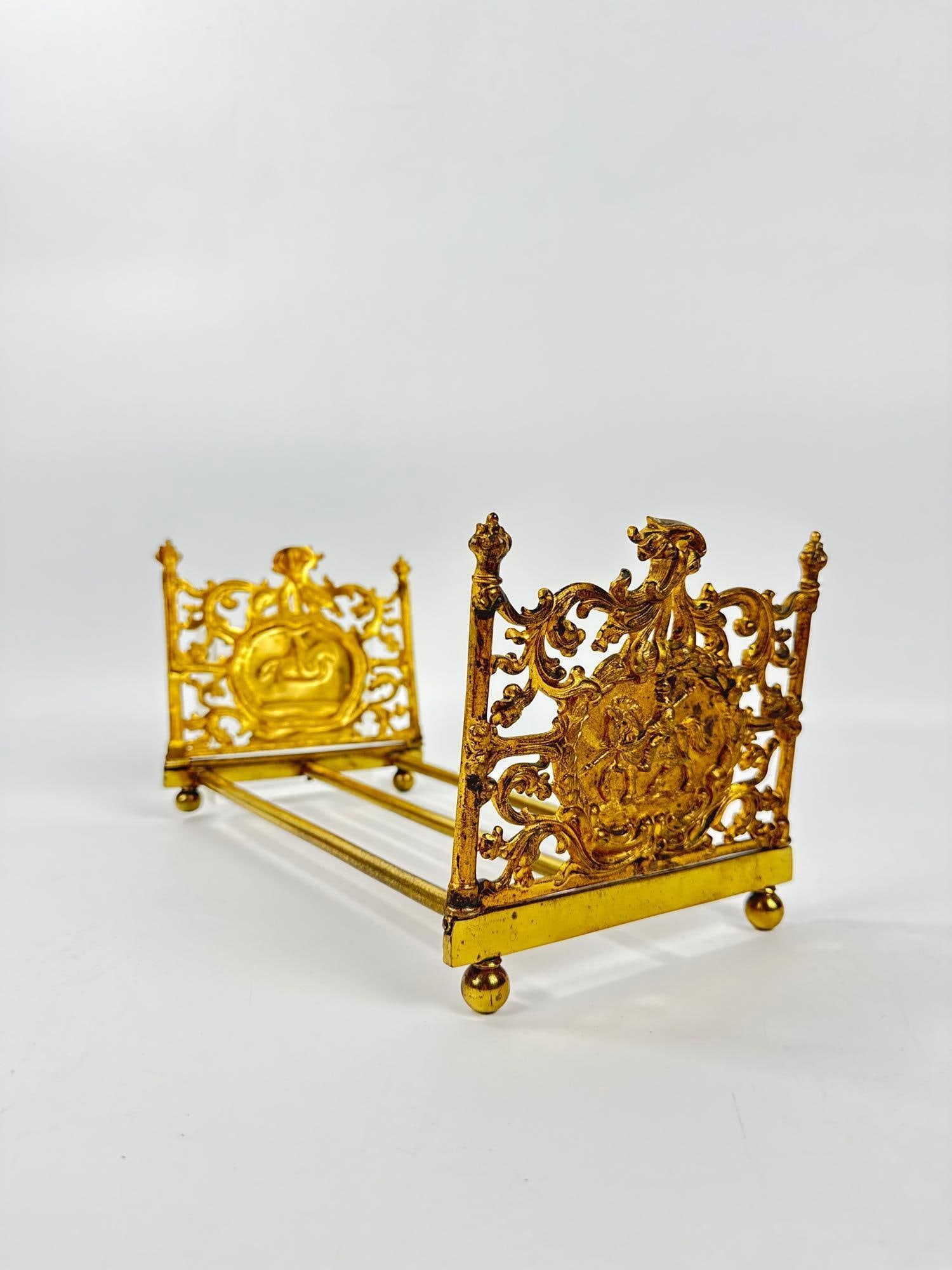 FRENCH BAROQUE STYLE ORNATE GILT BRASS EXPANDABLE BOOK SLIDE: Victorian era decorative desk accessory featuring pierced openwork end panels with classical crests and scrolling foliage on a sliding adjustable track. Approximate Dimensions: 6.5”h x 11&rdquo