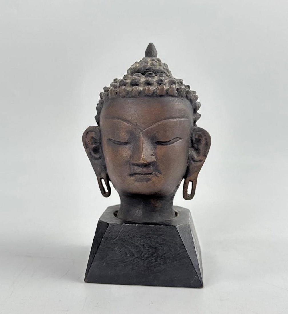THAI SUKHOTHAI STYLE CAST BRONZE BUDDHA HEAD: A petite, mounted bronze sculpture of a serene Buddha head with a traditional flame ushnisha finial. Approximate Dimensions: 6.74”H x 4.25”W x 3”D Artist or Maker: Unknown Provena