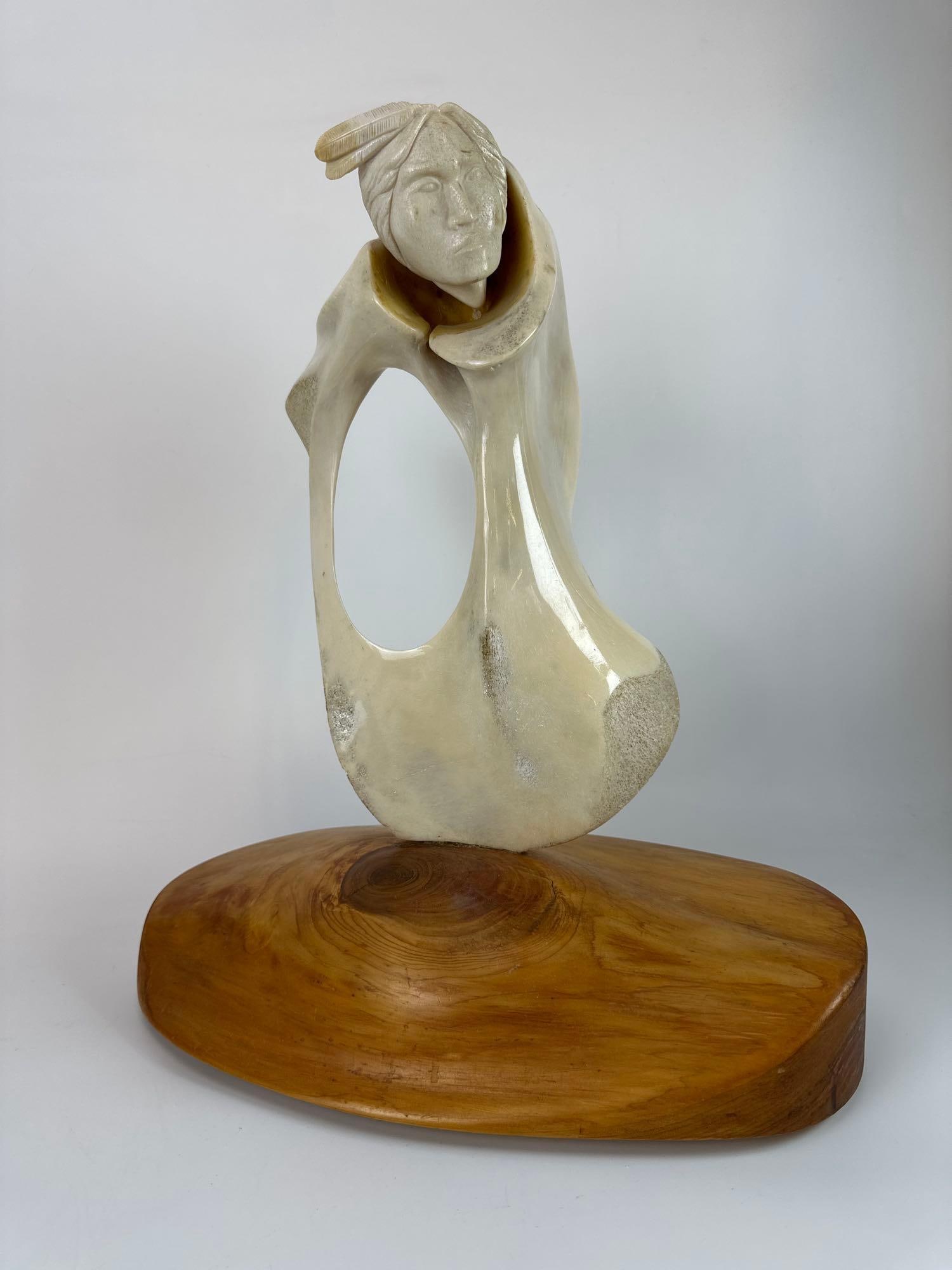 DIANE SCHENANDOAH ONEIDA CARVED SCULPTURE: Hand-carved organic caribou pelvis bone sculpture representing a Native American figure with a feathered headdress emerging from a stylized form on a polished wood plinth. Approximate Dimensions: App