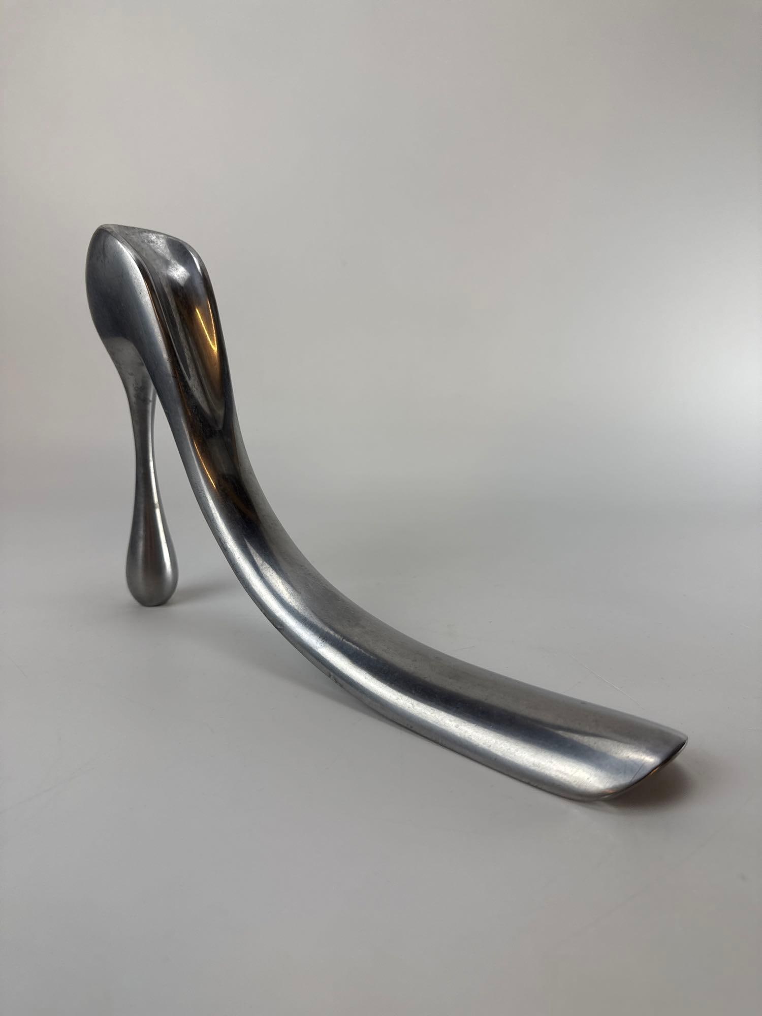SCULPTURAL CAST ALUMINUM STILETTO SHOE HORN BY MANOLO BLAHNIK: Sleek polished aluminum shoe horn in the form of an iconic stiletto heel designed for the 40th anniversary of Habitat. Approximate Dimensions: 11” x 2” x 7” h Artist or Maker: Manolo