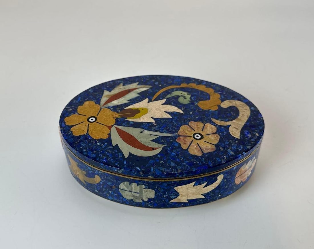 PIETRA DURA OVAL TRINKET BOX WITH LAPIS LAZULI AND SEMIPRECIOUS STONES: Oval lidded container composed of crushed lapis lazuli ground with floral and foliate pietra dura stone inlays. some losses. Approximate Dimensions: 1.75”H x 5.75”W x 4”D Artist or M