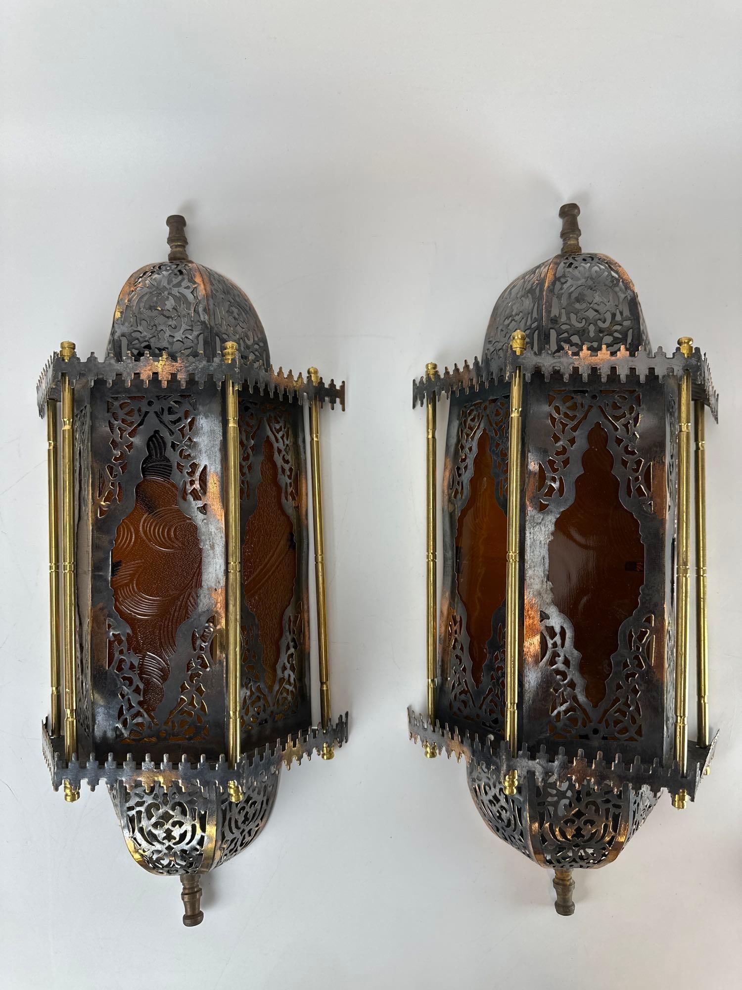 PAIR OF MOORISH REVIVAL PIERCED BRASS AND AMBER GLASS WALL SCONCES: Ornate Moroccan style lighting fixtures featuring intricate filigree metalwork and cylindrical amber-toned glass inserts. Approximate Dimensions: 20”x8”x5” Artist or Maker: Unknown