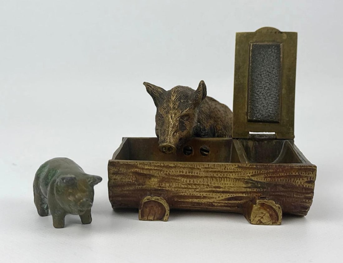 ANTIQUE AUSTRIAN BERGMAN TYPE BOAR VESTA WITH SOTHEBY’S PROVENANCE AND A CUTE PIGGY: A vintage brass wild boar vesta match safe and striker, featuring a Sotheby’s Arcade auction label dated April 7, 1982 (Lot #97-225). The piece is designed as a boar peering over a log-form match ho
