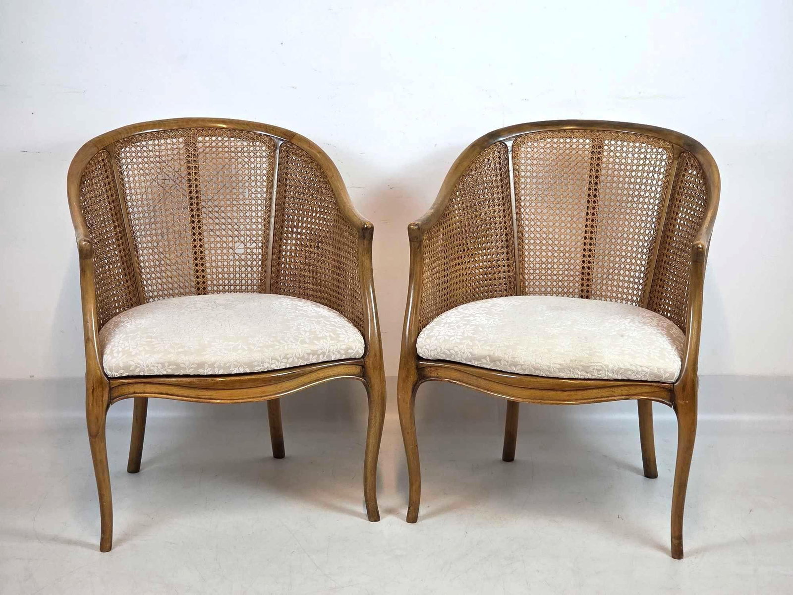 PAIR VINTAGE FRENCH PROVINCIAL CANE BACK BARREL CHAIRS IN THE MANNER OF BAKER FURNITURE OR MGEE & CO: A pair of French Provincial style carved wood barrel-back armchairs with woven caning panels, elegant cabriole front legs, splayed back legs, and upholstered brocade seats. Condition Note: There is mi
