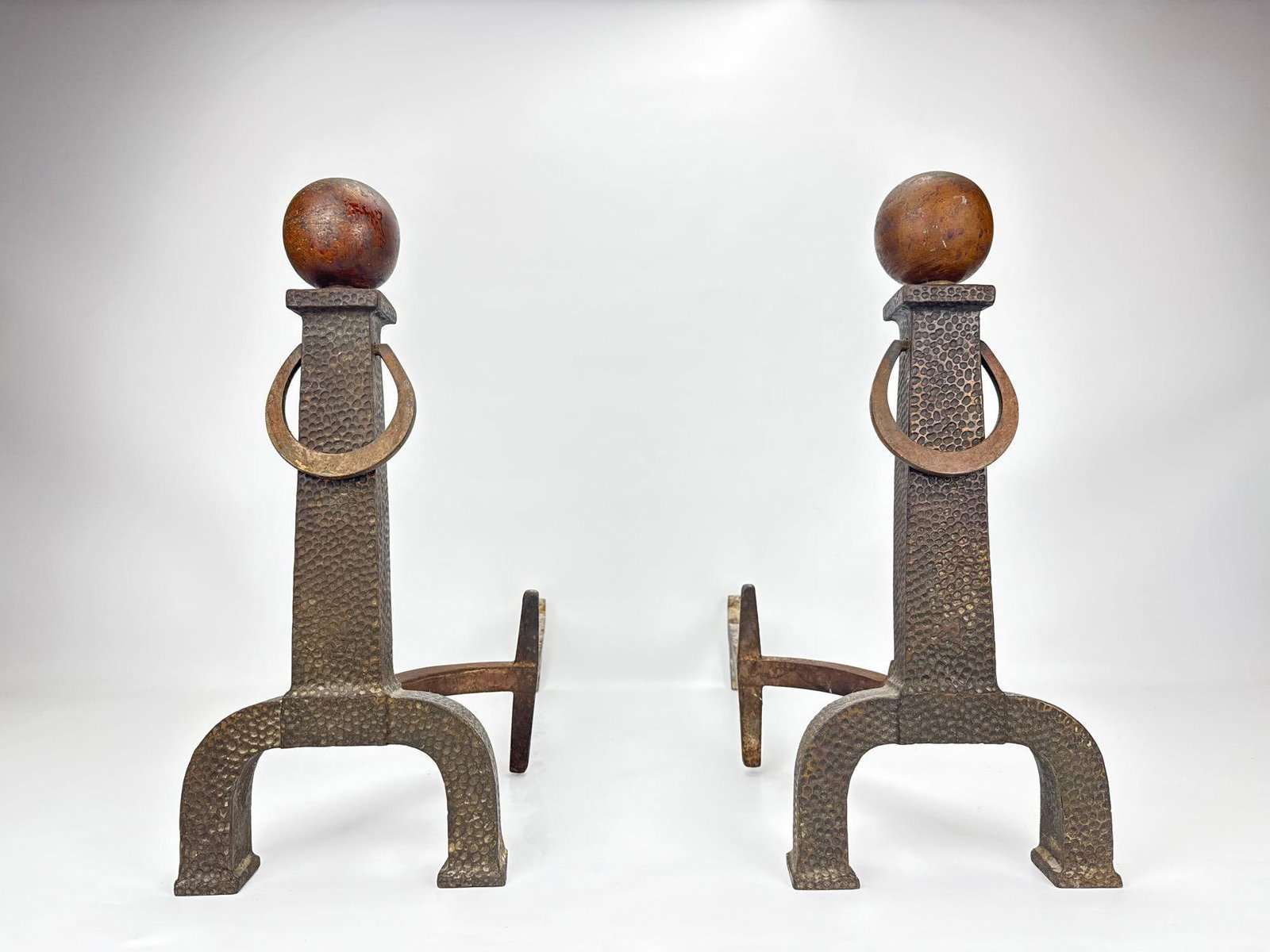 PAIR OF BRADLEY & HUBBARD HAMMERED ANDIRONS / CHENETS WITH BRASS BALL FINIALS AND RINGS: A pair of antique Arts & Crafts style hand-hammered iron fireplace andirons featuring a pitted textured finish, architectural arched bases, integrated ring handles, and turned wooden ball finials 