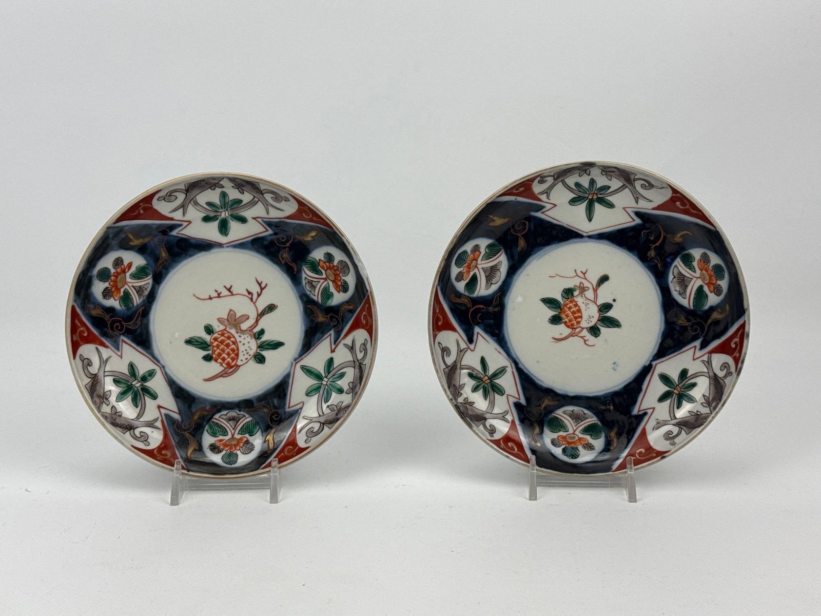 JAPANESE IMARI PORCELAIN SAUCERS PAIR: Pair of matching Japanese Imari hand-painted porcelain saucers decorated with a central pomegranate motif inside a geometric and floral paneled border Approximate Dimensions Larger Item: 1” H x 