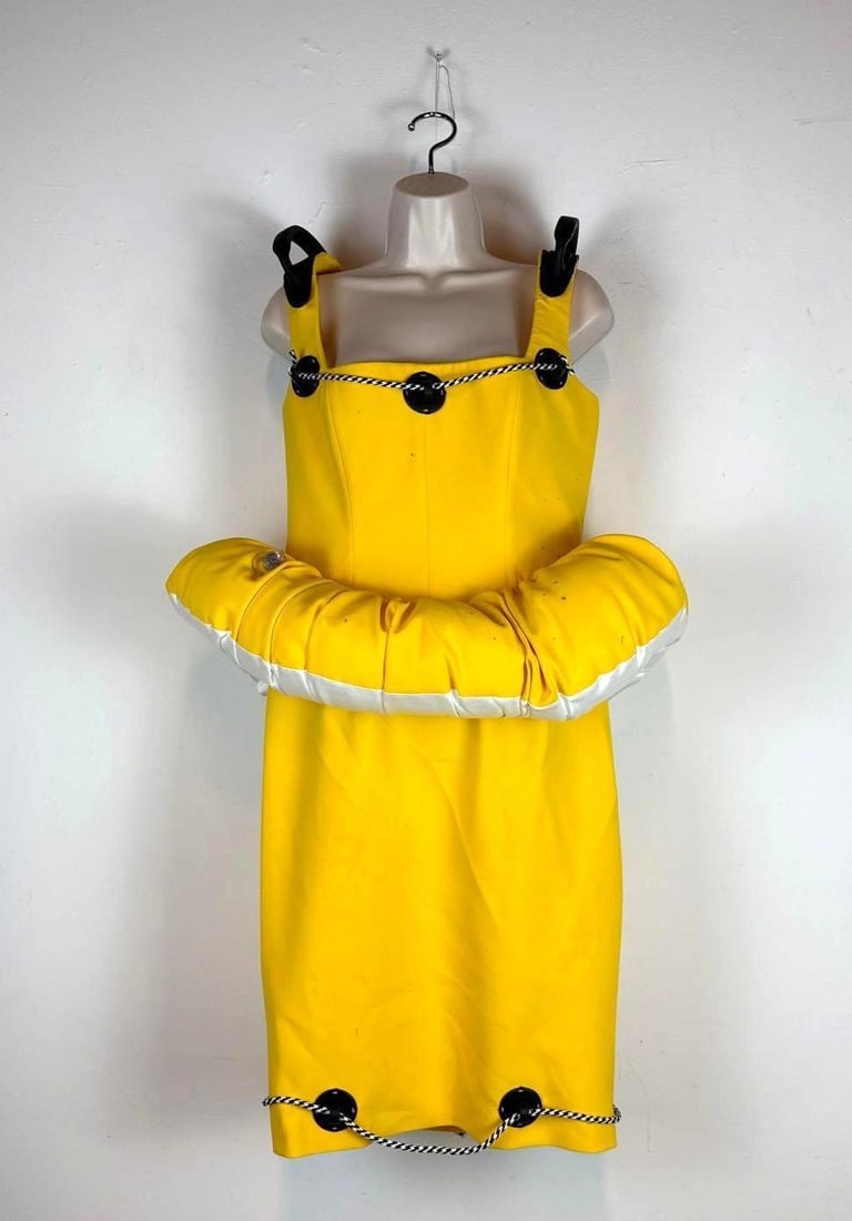 MOSCHINO COUTURE JEREMY SCOTT SPRING 2023 RUNWAY INFLATABLE LIFE RAFT DRESS: A bright yellow sleeveless midi-length sheath style dress with an integrated inflatable life raft waist ring and black-and-white braided safety cords along the neckline and hem. Please note splatter s