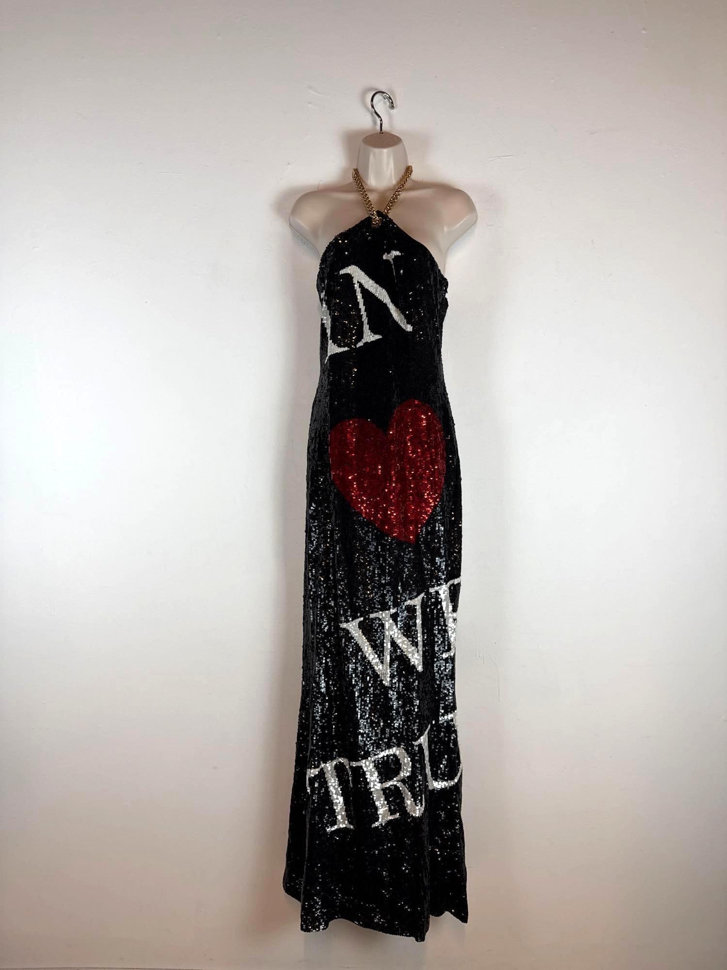 MOSCHINO COUTURE JEREMY SCOTT BLACK SEQUIN HALTER NECK "IN LOVE WE TRUST" GOWN (1 OF 2) SAMPLE: A full-length halter neck dress entirely embellished in shimmering black sequins with a chunky gold-tone chain strap, a large central red sequin heart motif, and white sequin "IN LOVE WE TRUST"