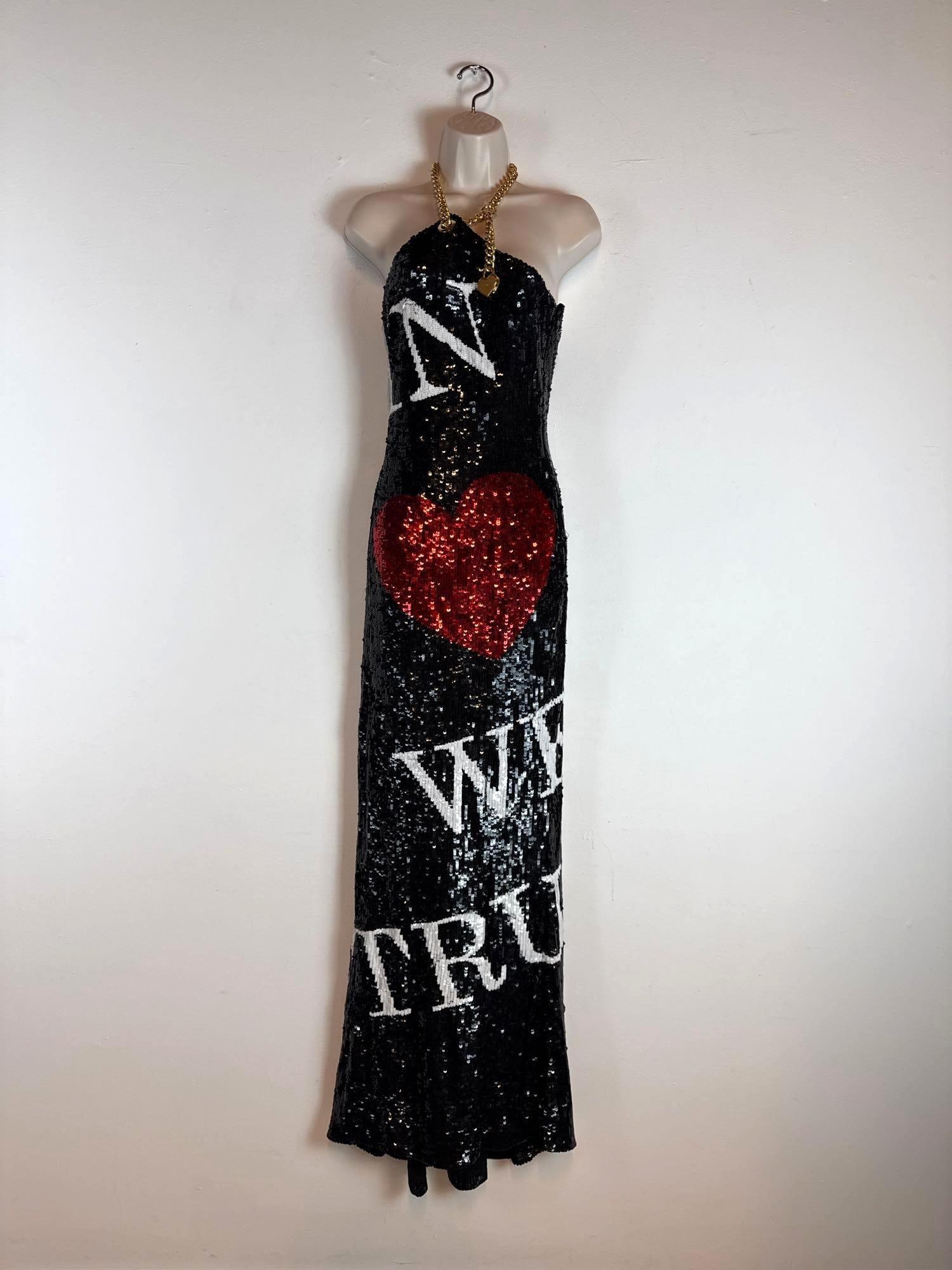 MOSCHINO COUTURE JEREMY SCOTT BLACK SEQUIN HALTER NECK "IN LOVE WE TRUST" GOWN (2 OF 2) SAMPLE: A full-length halter neck dress entirely embellished in shimmering black sequins with a chunky gold-tone chain strap, a large central red sequin heart motif, and white sequin "IN LOVE WE TRUST"