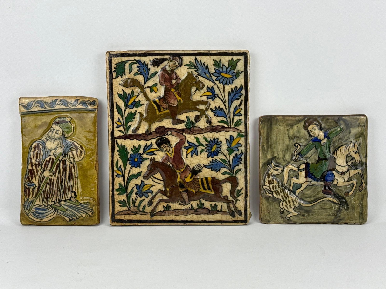 COLLECTION OF THREE PERSIAN QAJAR DYNASTY CERAMIC TILES: Group of three 19th-century Persian Qajar molded pottery polychrome glazed cermaic architectural tiles of equestrian hunters, and a seated elder with floral border. Approximate Dimensions: Largest 12.