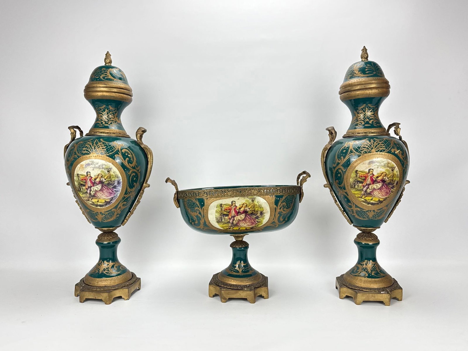 SEVRES STYLE PORCELAIN AND ORMOLU GARNITURE SET OF 3: Antique French style teal green-ground porcelain three-piece mantle set consisting of two lidded urns and a central pedestal bowl decorated with hand-painted romantic couple vignettes and ornate gilt 