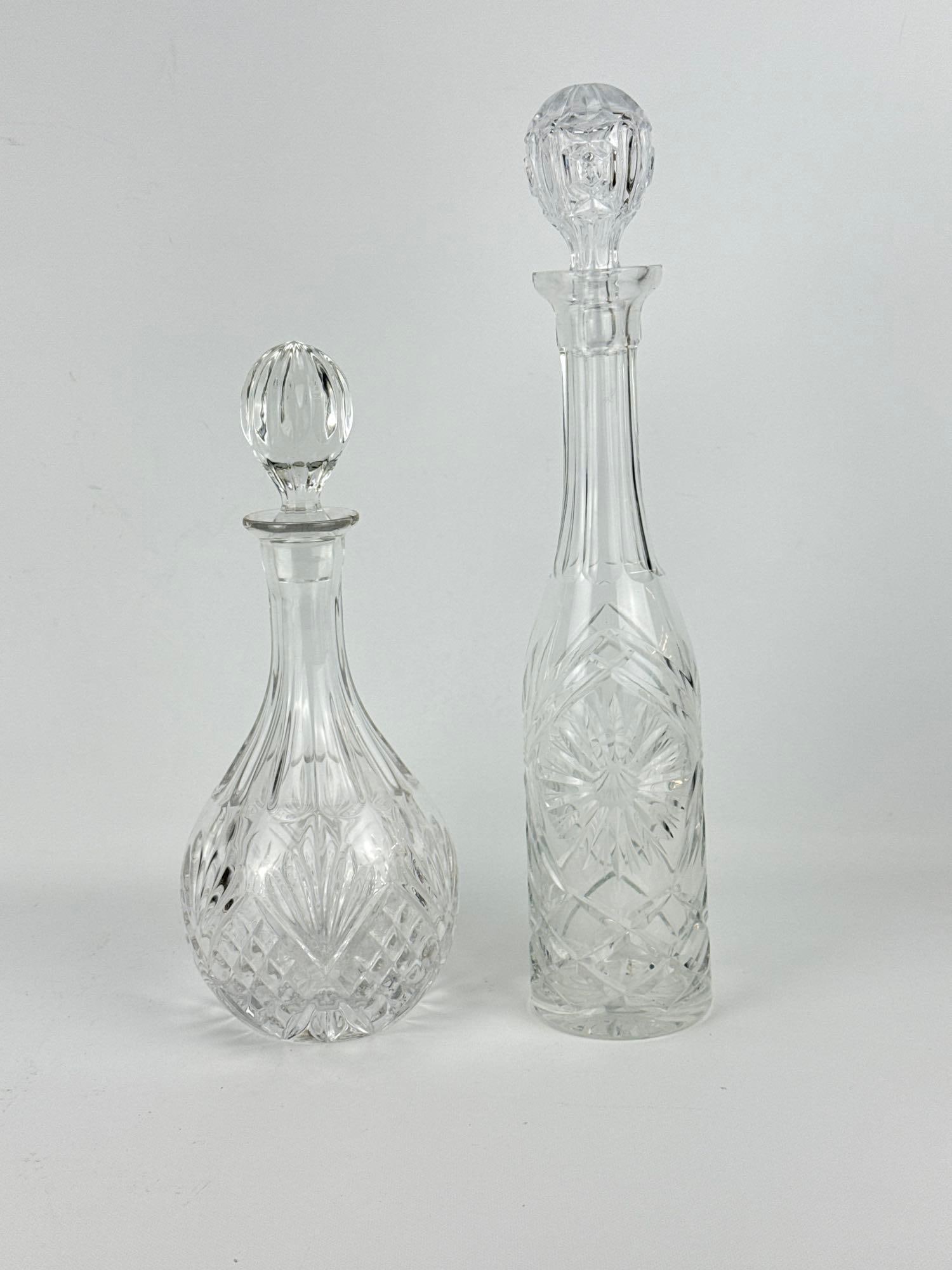 TWO CUT CRYSTAL GLASS DECANTERS WITH STOPPERS: *An assortment of two clear cut crystal decanters including a tall cylindrical bottle decanter with starburst panels and a classic bulbous spirit decanter, both wit faceted glass stoppers and geometri