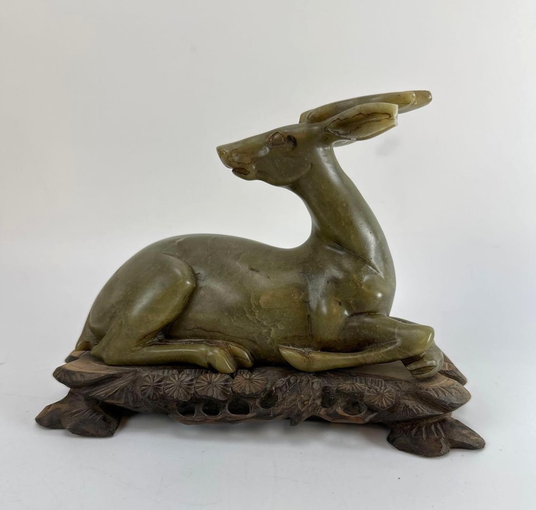 CHINESE HARDSTONE / JADEITE CARVED RECUMBENT DEER ON STAND: *20th-century Chinese carved hardstone (nephrite or bowenite) figure of a deer on a custom-fitted pierced openwork hardwood display stand. Approximate Dimensions: 8"x7x3” Artist or Maker: Un