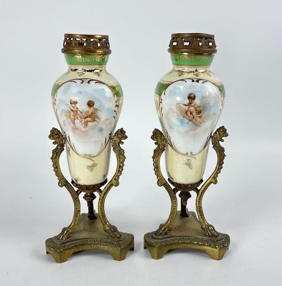 PAIR OF FRENCH PORCELAIN SEVRES STYLE URNS: *A pair of petite, vase-shaped porcelain urns with a creamy white and pale green ground, decorated with hand-painted panels depicting putti or cherubs amidst clouds, each mounted in a patinated bronze