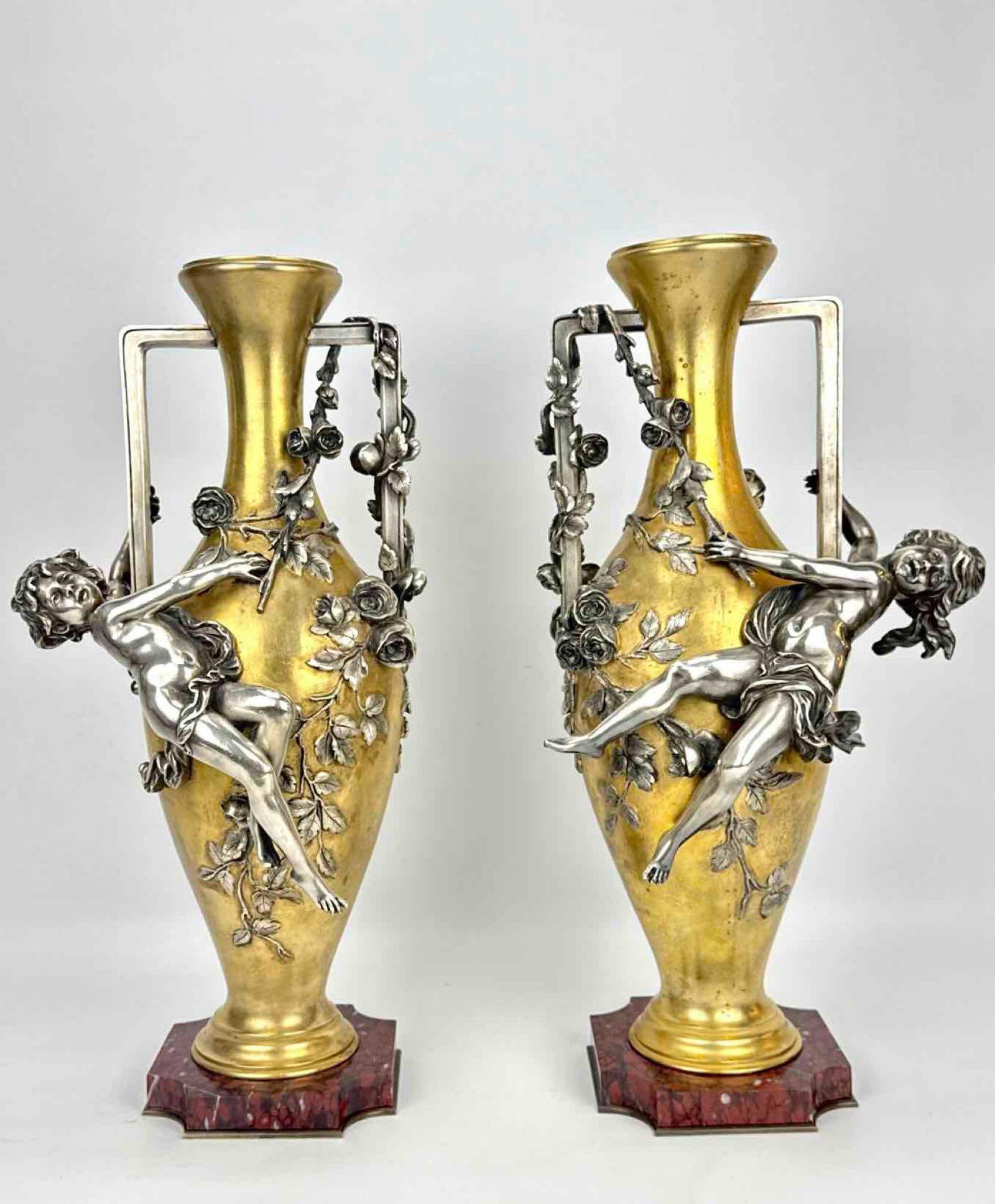 AUGUSTE MOREAU (FRENCH, 1834-1917), PAIR OF BRONZE FIGURAL VASES: *Pair of 19th-century French silvered and gilt metal amphora-form vases rouge griotte marble bases with high-relief sculpted putti scaling rose vines. Approximate Dimensions: 21.75"H x 10"w x 