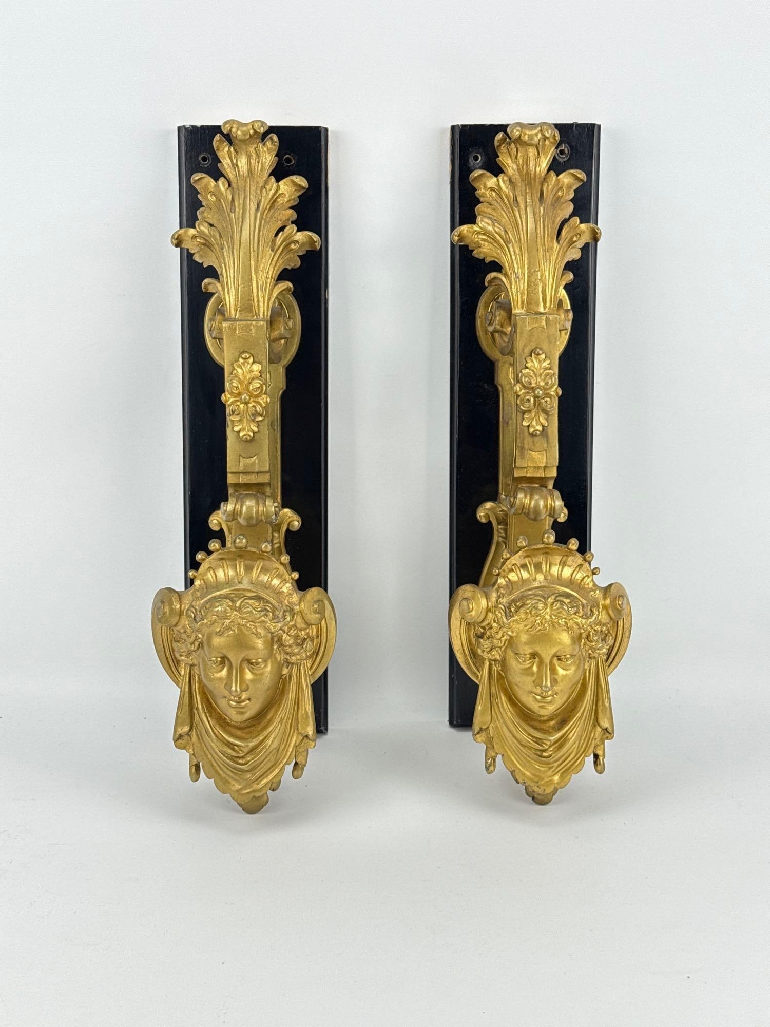 PAIR OF MONUMENTAL FRENCH NEOCLASSICAL GILT BRONZE ORMOLU MASK DOOR PULLS: Pair of large-scale architectural gilt bronze door handles depicting classical female masks mounted on ebonized wood backings. Approximate Dimensions: 18” H X 4.75” W X 8” D Artist o