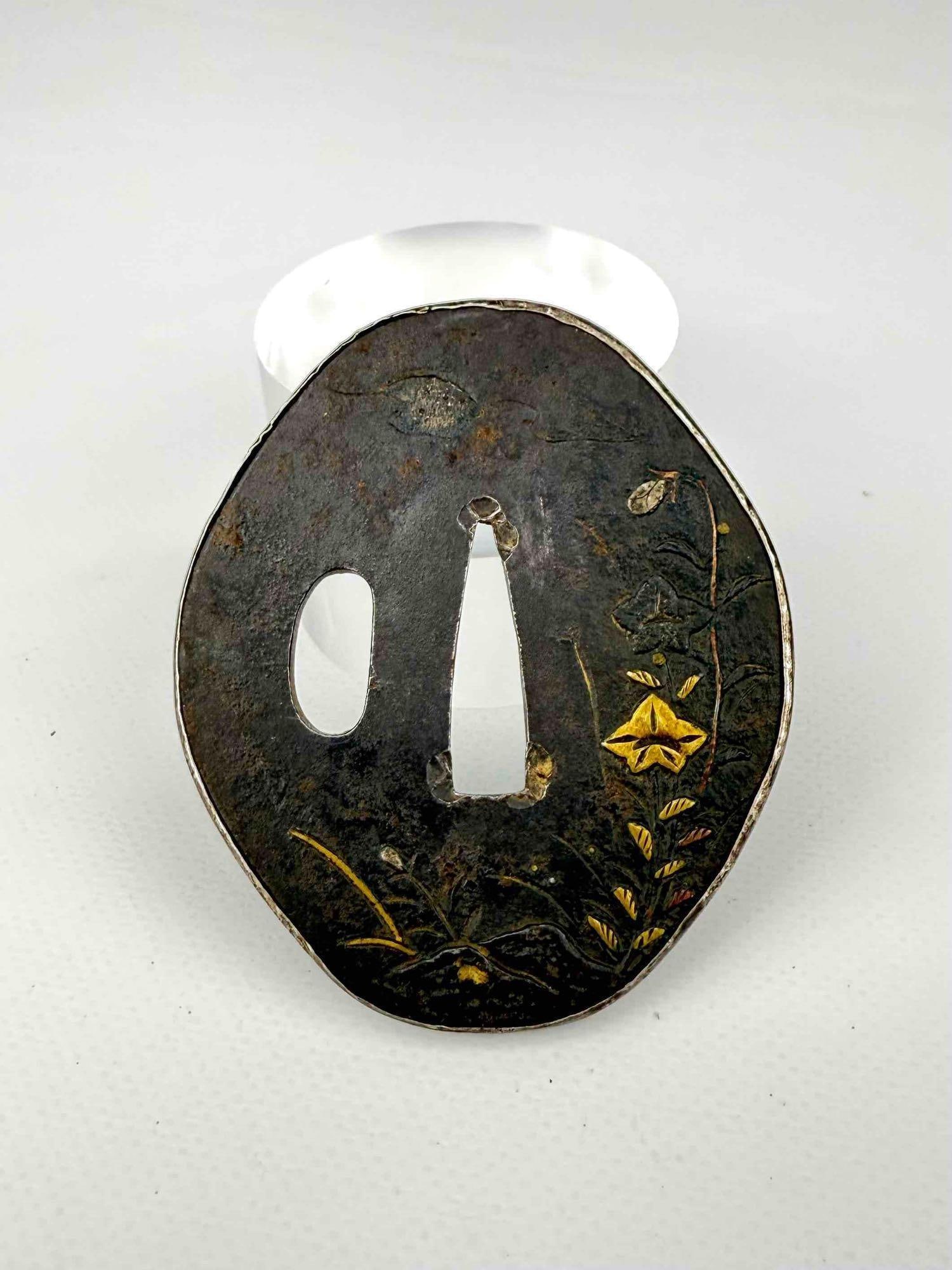 JAPANESE ANTIQUE GOLD INLAY SHAKUDO OR BRONZE SUBA SWORD GUARD: An 18th to 19th-century Edo period oval-shaped patinated bronze or shakudo tsuba embellished with delicate mixed metal and gold inlay detailing depicting wild floral blooms, autumn grasses, and a flyi