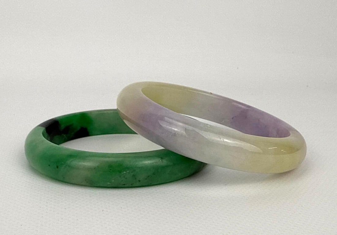 CHINESE ANTIQUE NATURAL LAVENDER AND EMERALD GREEN JADEITE BANGLE BRACELET PAIR: A pair of two genuine natural jadeite jade continuous bangle bracelets, including one vibrant spinach or emerald green stone with darker inclusions and one multi-toned piece displaying soft lavender v