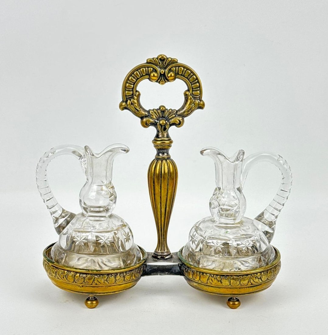 VICTORIAN BACCARAT STYLE CUT GLASS CRUET DECANTER SET IN BRASS STAND: Two starburst cut crystal oil and vinegar cruets with applied handles housed in an ornate footed brass caddy with a central ring handle Approximate Dimensions: 6.5” H X 9” W X 4” D A