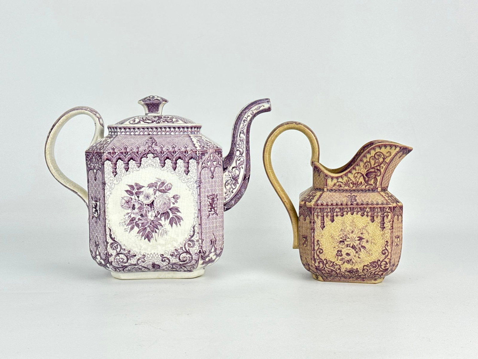 ANTIQUE GERMAN VILLEROY & BOCH METTLACH STEINGUT PURPLE TRANSFERWARE TEAPOT AND PITCHER: Two-piece assembly of octagonal neo-gothic ironstone hollowware including a covered teapot marked Villeroy & Boch Mettlach and a cream pitcher decorated with purple floral bouquets and heraldic cr