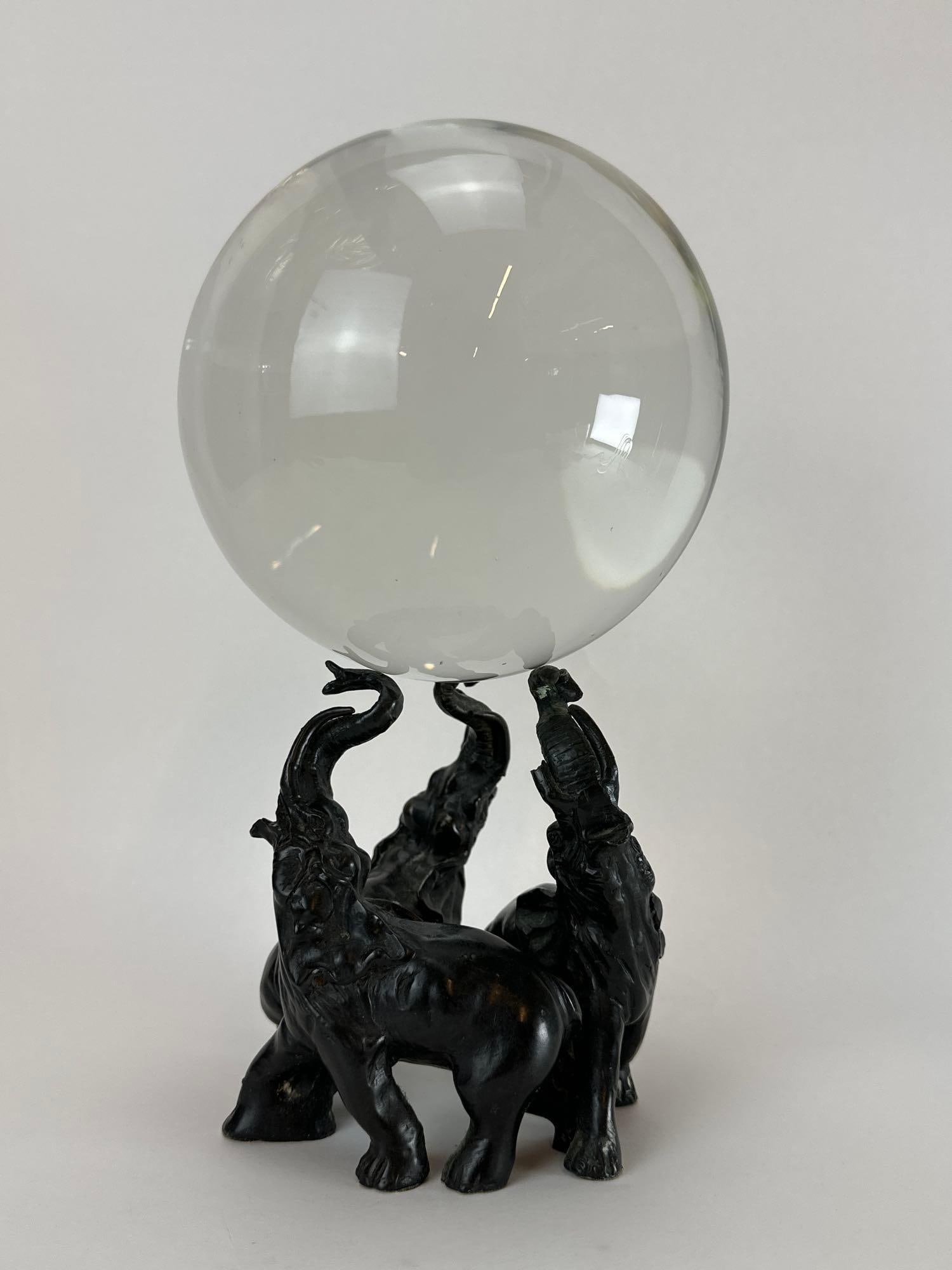 HOLLYWOOD REGENCY BACCARAT STYLE ORB ON PATINATED METAL THREE ELEPHANTS BASE PEDESTAL: Polished clear glass or crystal sphere balancing atop three standing patinated cast metal elephants with raised trunks. Approximate Dimensions: 10” H x 6” Dia. Artist or Maker: Unknown Pro