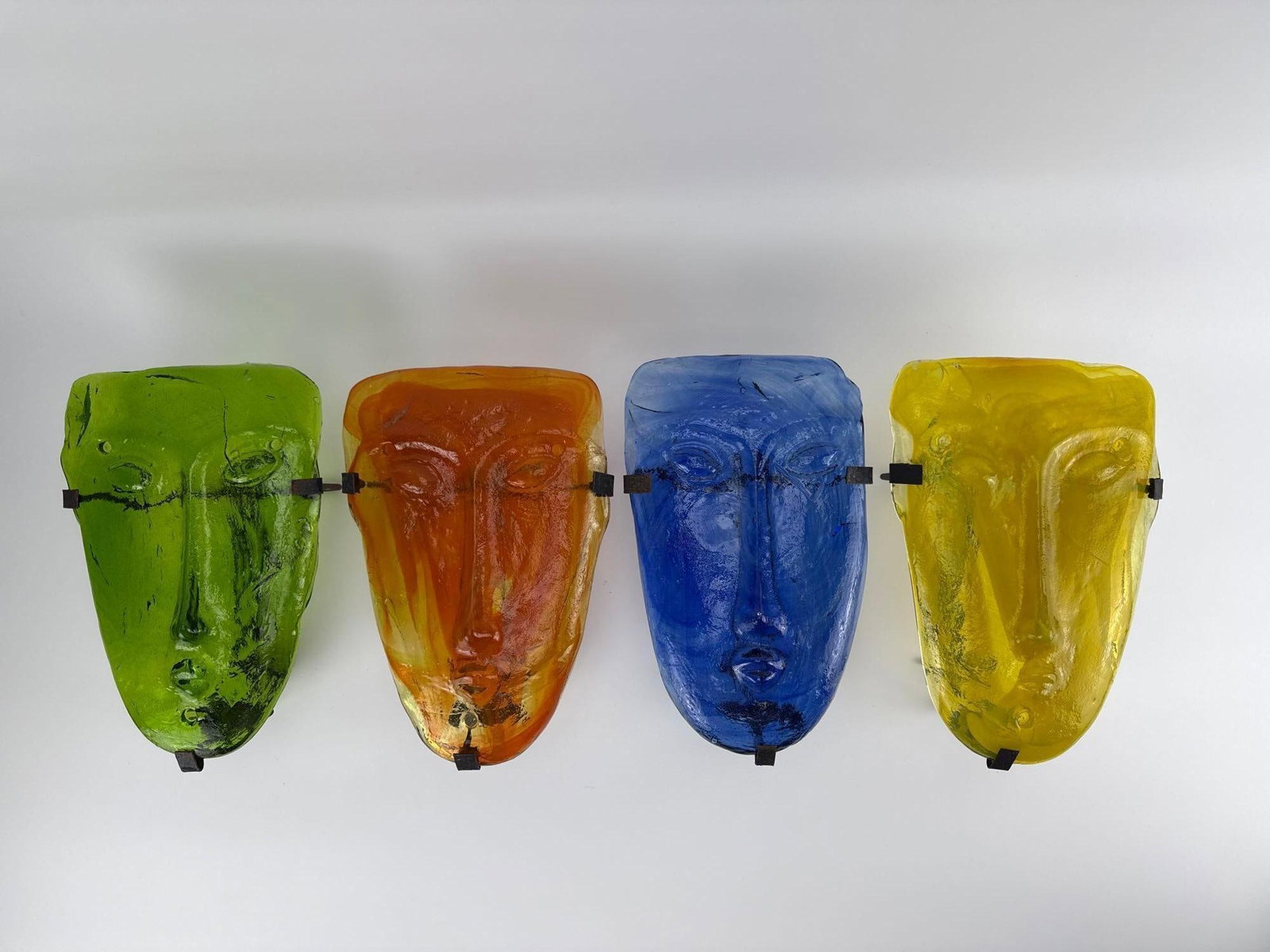 FOUR VINTAGE SCANDINAVIAN MODERN GLASS FACE MASK CANDLE SCONCES IN THE MANNER OF ERIK HOGLUND: Set of four 1960s mouth-blown amber, blue, yellow, and green art glass face mask candle sconces mounted in black metal wall brackets. Approximate Dimensions: 11" X 7" X 5" DEEP Artist or M