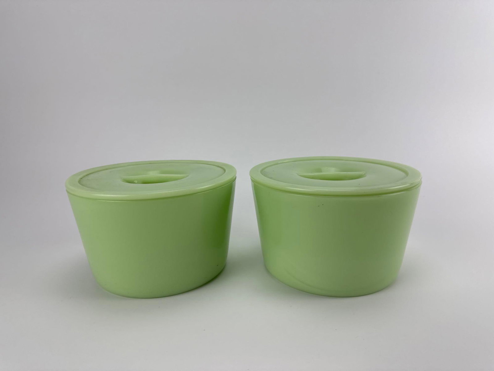 PAIR OF VINTAGE JEANNETTE GLASS CO. JADEITE ROUND REFRIGERATOR STORAGE CROCKS WITH LIDS: Pair of antique 1930s opaque green uranium jadeite glass round leftovers refrigerator crocks with matching flat tab-handled lids. Approximate Dimensions: 3.75” x 6” Dia Artist or Maker: Je