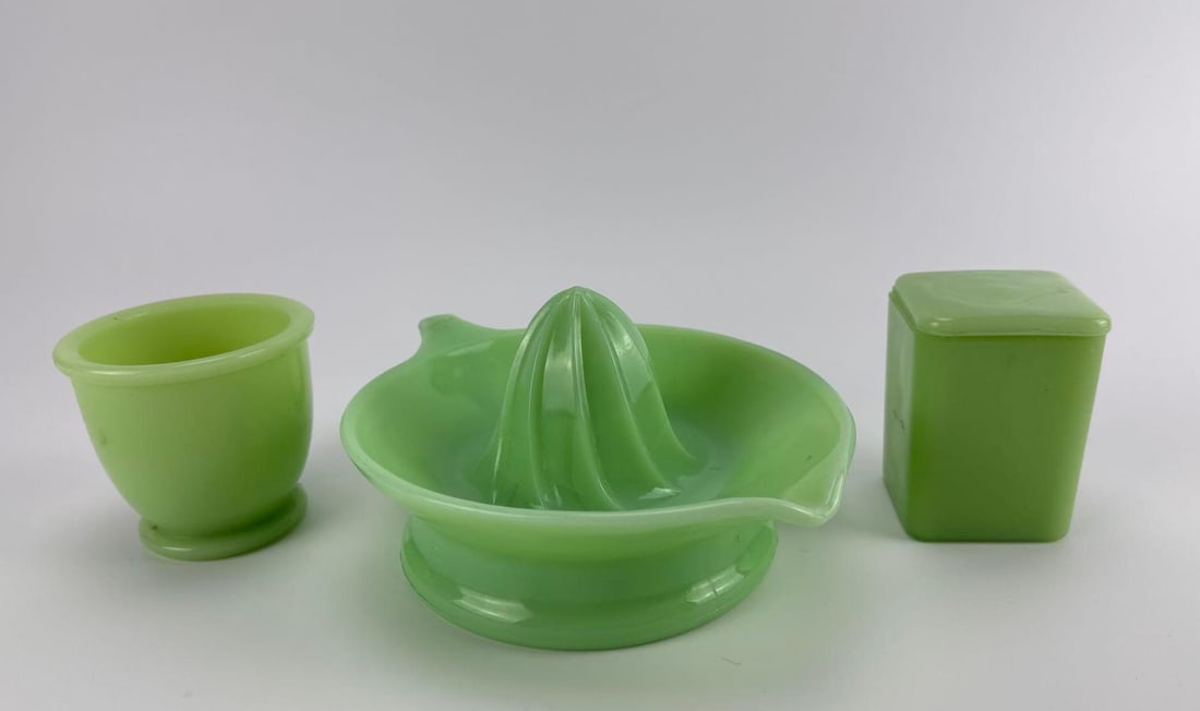 THREE VINTAGE JEANNETTE AND MCKEE GLASS CO. JADEITE KITCHENWARE COLLECTIBLES INCLUDING JUICER: Three-piece antique 1930s opaque green uranium jadeite glass kitchen set comprising a citrus juicer reamer, a small square lidded spice container, and a footed juice cup. Approximate Dimensions: