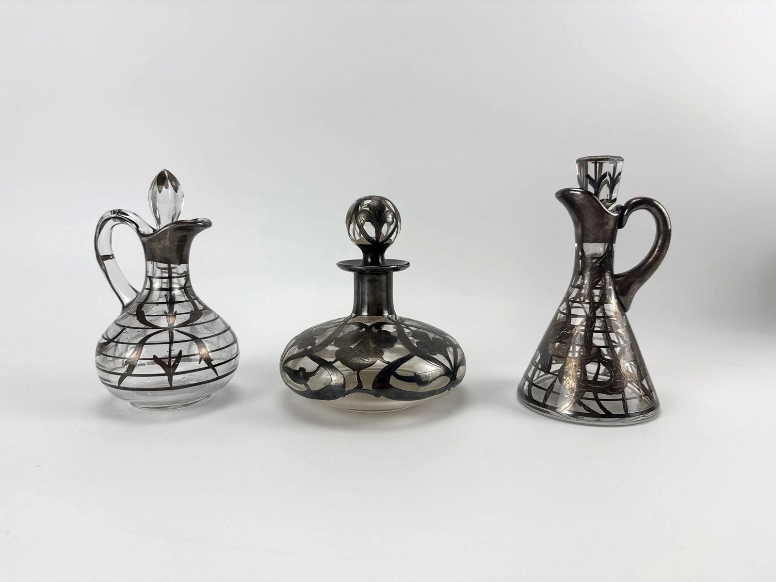LOT OF SILVER-OVERLAY HAND-BLOWN GLASS CRUETS AND PERFUME BOTTLE WITH STOPPERS: Group of hand-blown glass cruets and a perfume bottle with silver overlay decoration and stoppers. Note chip to stopper base in photo. Approximate Dimensions Tallest: 6.5" H X 3.5" Dia. Artist