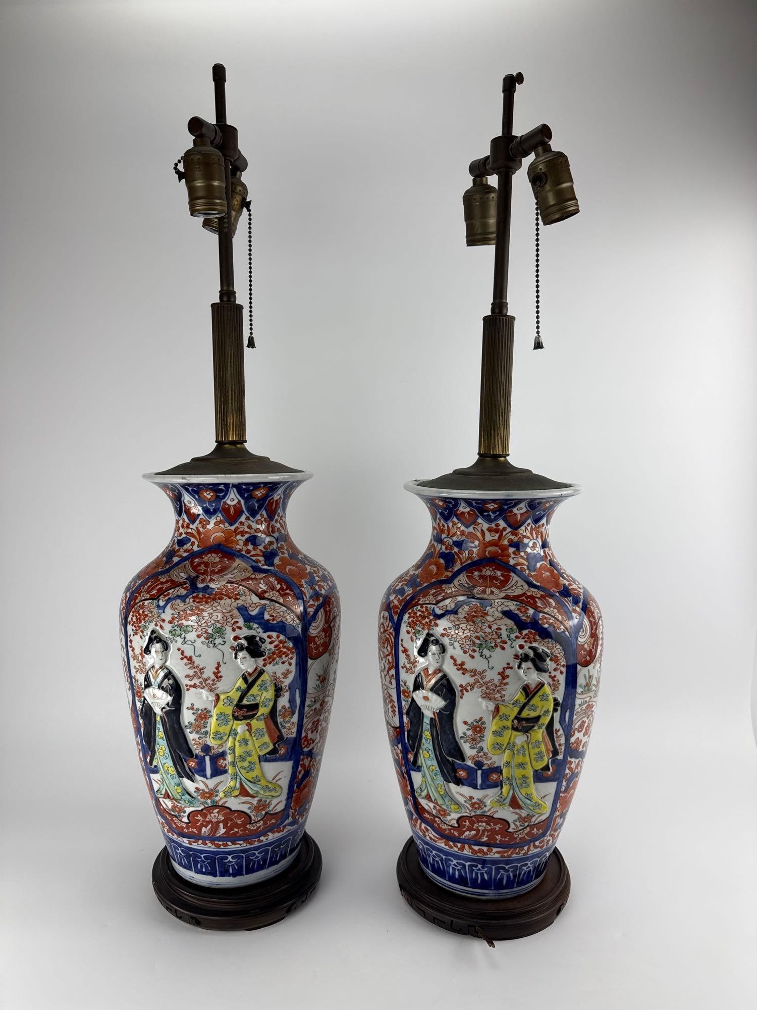 PAIR JAPANESE IMARI HAND PAINTED PORCELAIN VASE TABLE LAMPS: *Pair of Japanese Imari porcelain baluster vases hand-painted with geisha cartouches mounted as table lamps on carved wood bases. Approximate Dimensions: 8” dia x 31” T Artist or Maker: Un