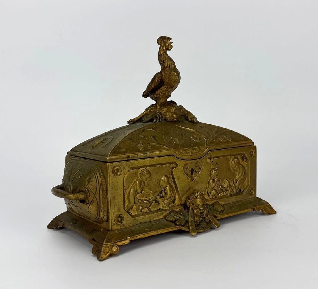 ANTIQUE FRENCH RENAISSANCE REVIVAL BRASS CASKET WITH ROOSTER FINIAL: *A charming nineteenth-century ornate cast brass jewelry box or vanity casket relief-decorated with scenes of classical playful putti, complete with side loop handles, a masqueron footed base, and a p