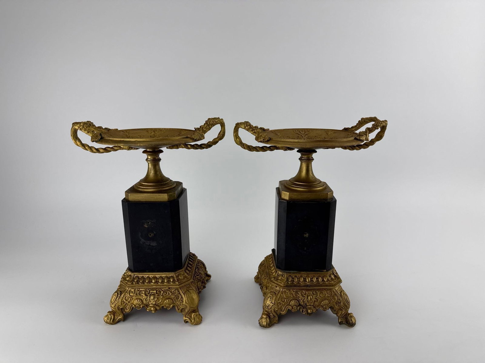 PAIR FRENCH FRENCH NEOCLASSICAL GILT BRONZE ORMOLU AND BLACK MARBLE TAZZAS: *Pair of 19th-century French Neoclassical gilt bronze shallow tazza dishes with scrolled handles on black marble pedestals over ornate cast bases. Approximate Dimensions: 7.5” x 5.5” x 10&
