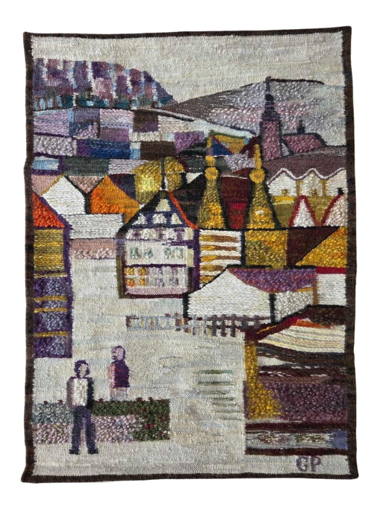 HAND LOOMED WOOL TAPESTRY "MOUNTAINSIDE VILLAGE": Hand-loomed wool tapestry / wall hanging depicting a stylized mountainside village of half-timbered houses with a church spire and figures, worked in purple, orange, gold and cream tones; woven with a