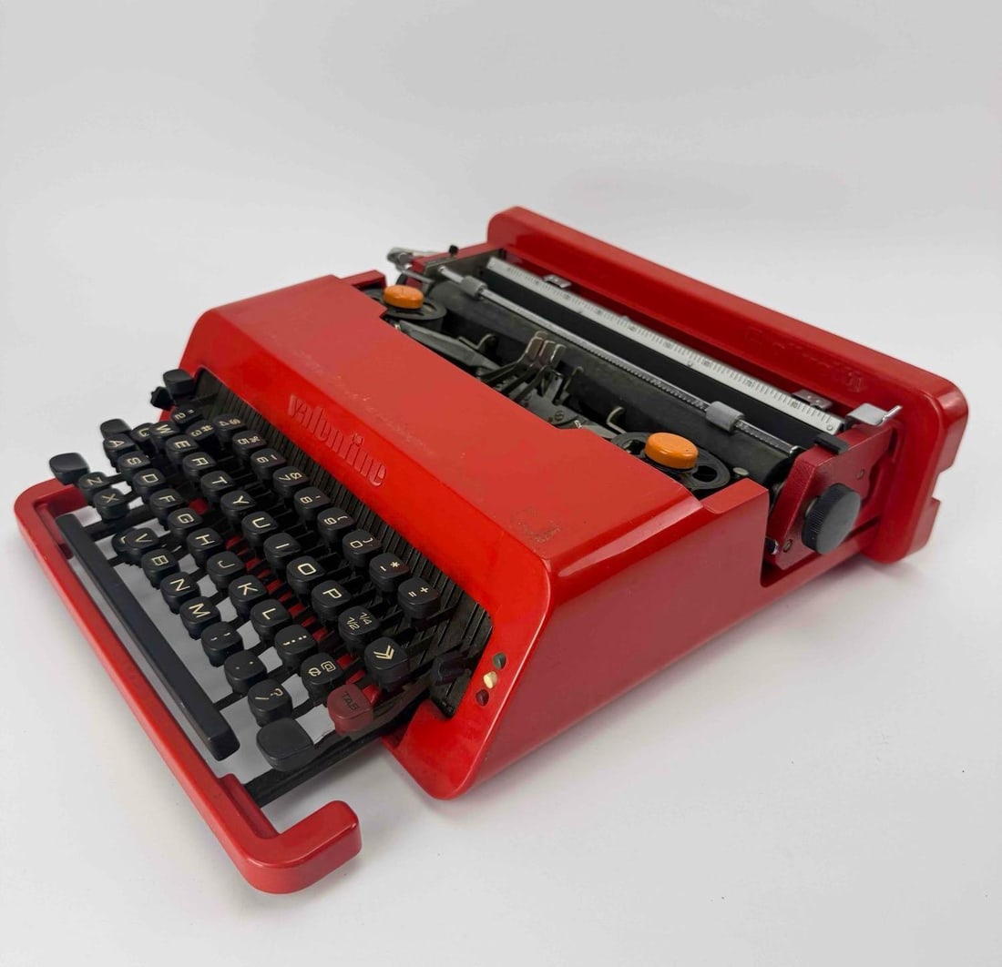 OLIVETTI VALENTINE RED PORTABLE TYPEWRITER, ETTORE SOTTSASS DESIGN, WITH CASE: Red plastic portable typewriter with matching snap-on carrying case, orange spool caps and original printed advertising tag. Approximate Dimensions: 4.5" H x 13.5" W x 14" D Artist or Make