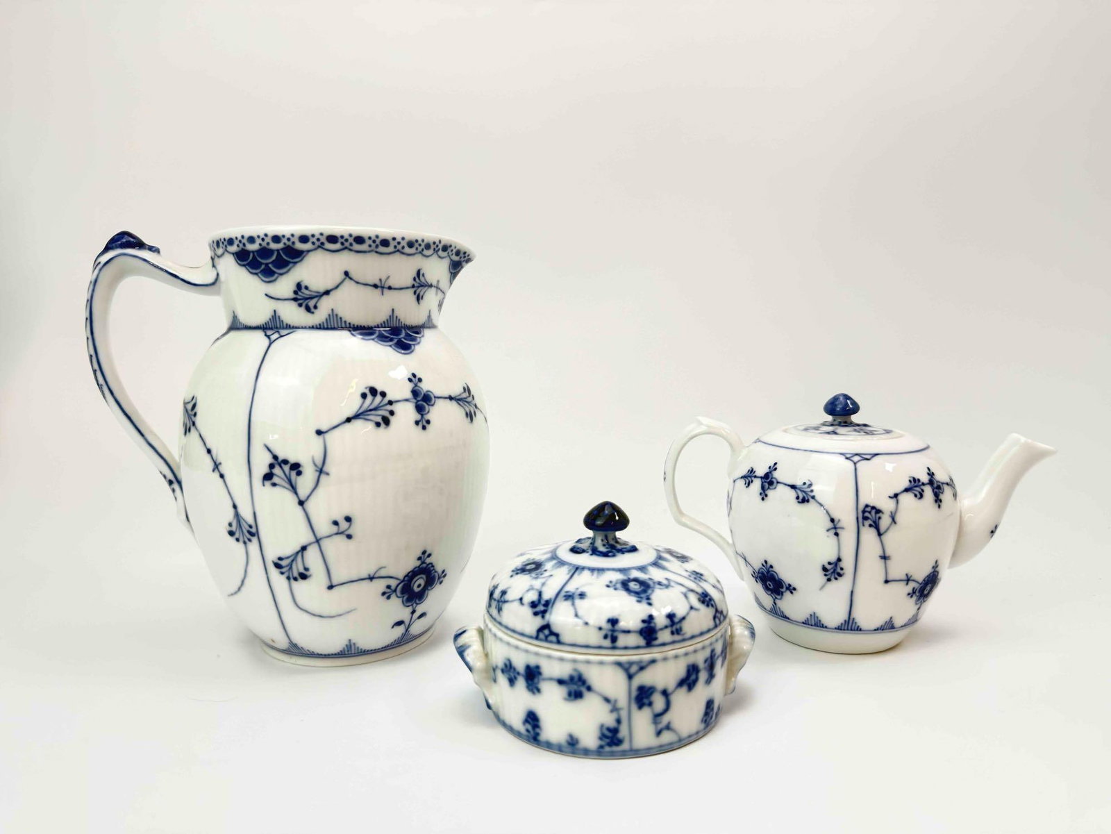 ROYAL COPENHAGEN BLUE FLUTED HALF LACE PORCELAIN PITCHER TEAPOT & SUGAR BOWL TRIO: A 3-piece, hand-painted blue and white porcelain table service including a pitcher, small teapot, and covered sugar bowl. Approximate Dimensions: 6.75” H x 6” W Artist or Maker: Royal Cope