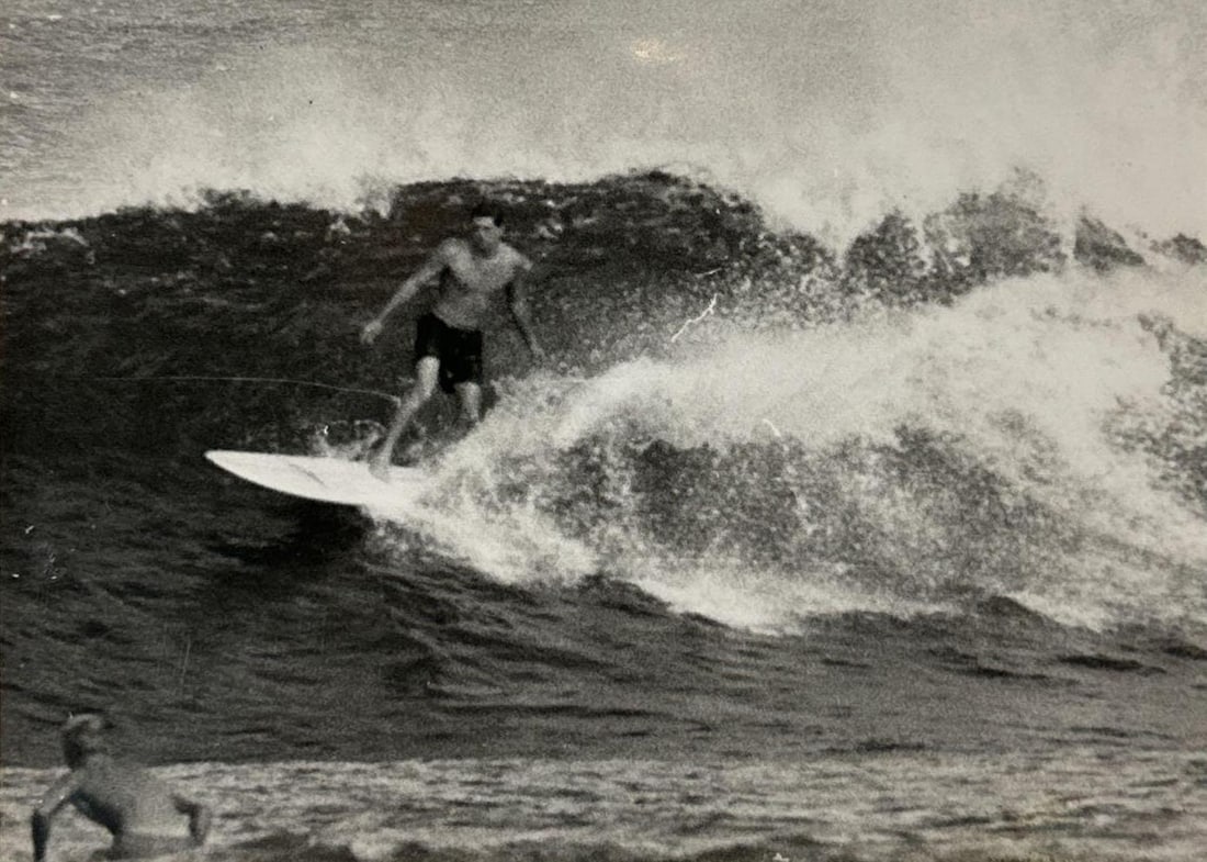 VAL VALENTINE VINTAGE SURF PHOTOGRAPH OF BUTCH VAN ARTSDALEN, HALEIWA 1963: Vintage black-and-white surf photograph of a longboard surfer trimming across a North Shore wave, a second surfer swimming in the foreground. Approximate Dimensions: Frame: 20"H x 24"W Artist