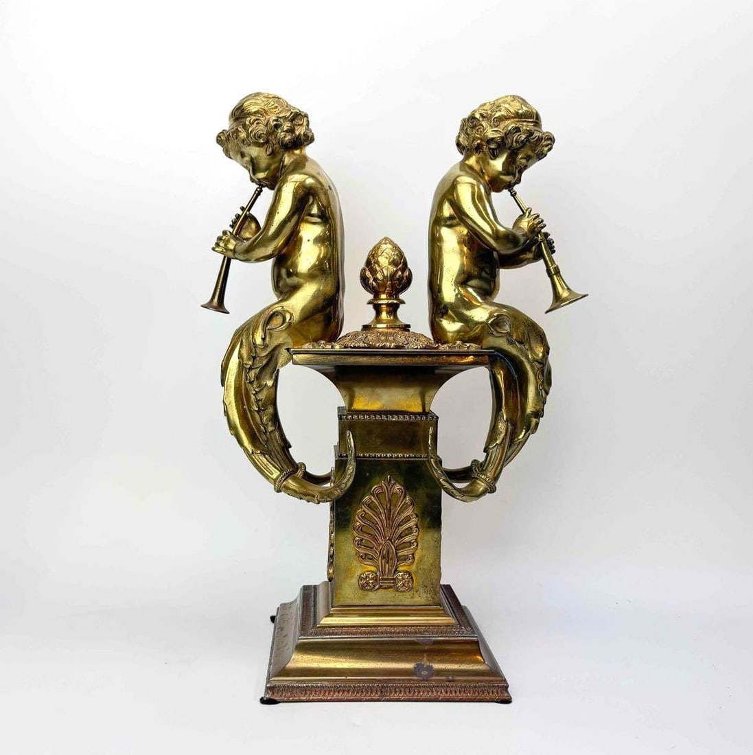 GILT BRASS FIGURAL GROUP OF TWO TRUMPETING PUTTI ON A CLASSICAL PEDESTAL: Figural group in gilt brass, two winged putti blowing trumpets seated back to back above a tapering pedestal cast with anthemion and mask ornament, on a stepped square base with ball feet Approximate 